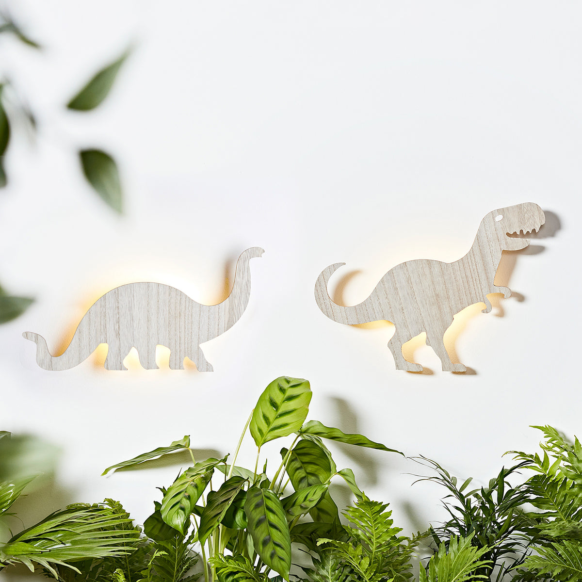 Zwei Dinosaurier Wandlampen aus Holz, Diplodocus und T-Rex, beleuchtet an weißer Wand mit grünen Pflanzen