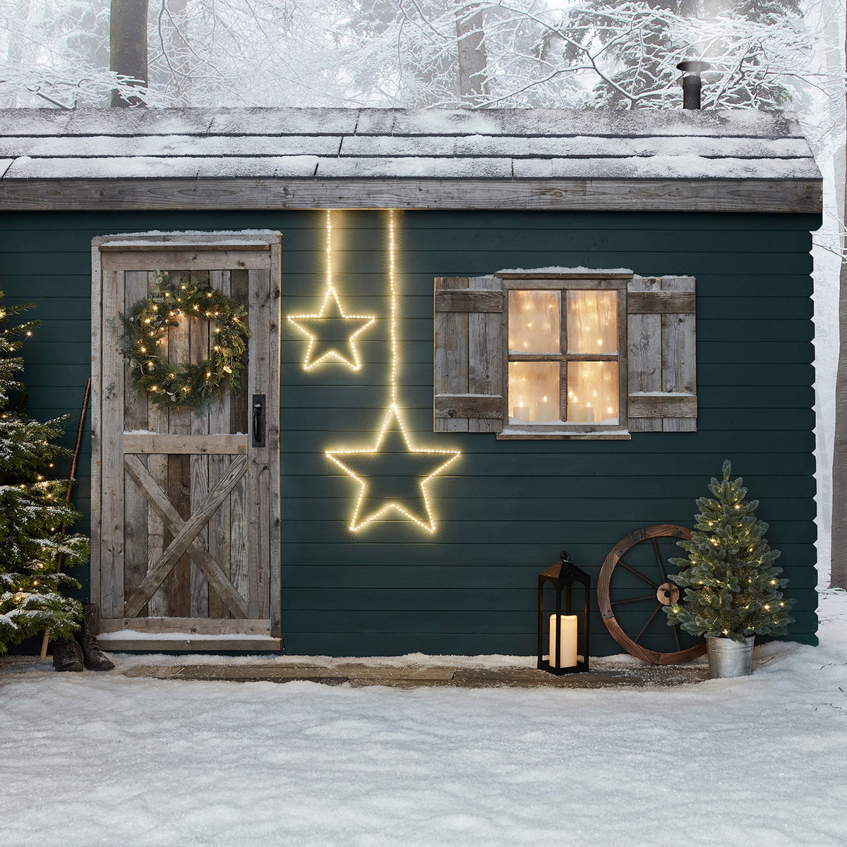Zwei leuchtende LED-Sterne an einer grünen Hauswand mit Tür, Fenster und winterlicher Dekoration im Schnee