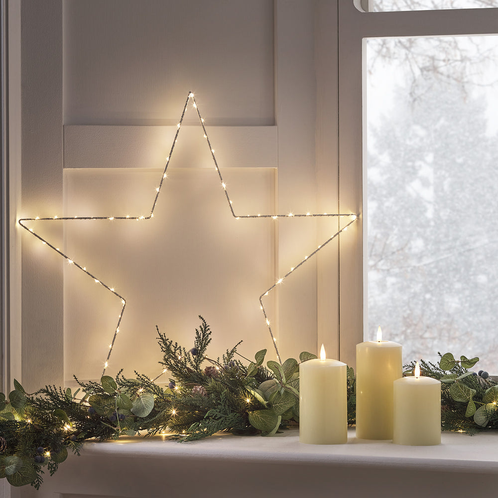 Beleuchteter LED-Stern an weißer Wand über Fensterbank mit grüner Beerengirlande und drei elfenbeinfarbenen LED-Kerzen