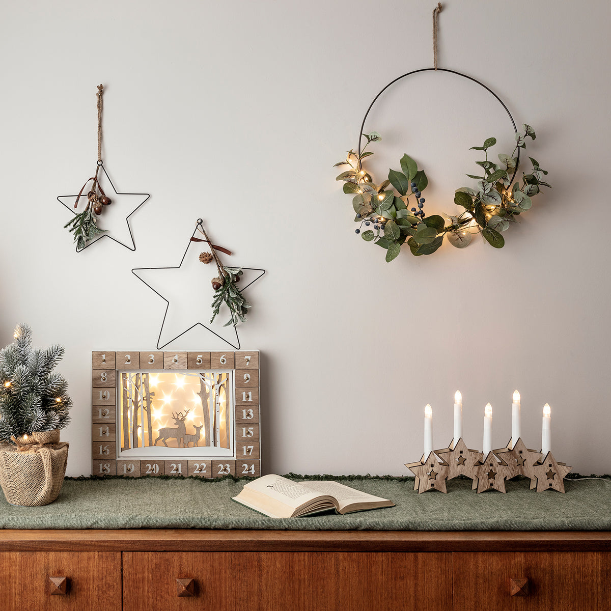 Weihnachtskranz mit Lichterkette an Wand neben Adventskalender, Kerzen und Stern-Deko auf Sideboard