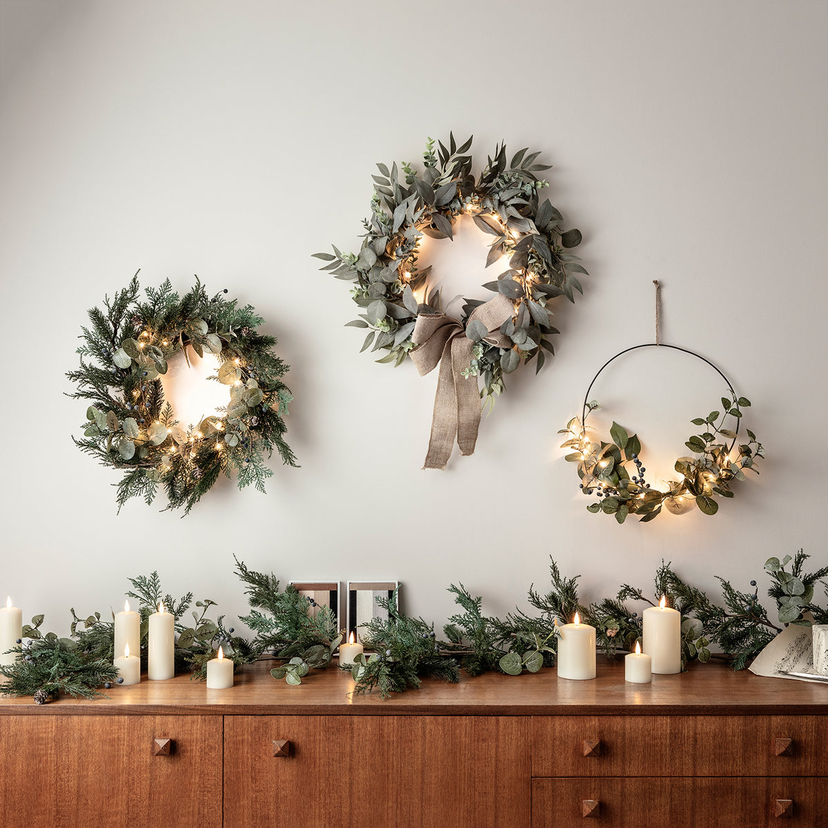 Drei verschiedene Weihnachtskränze mit Lichterketten an Wand über Sideboard mit Kerzen und Tannengrün