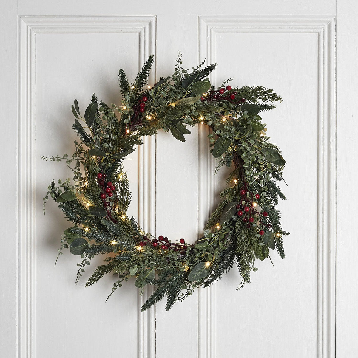 Weihnachtskranz mit grünen Zweigen, roten Beeren und Micro LED Lichterkette an weißer Tür