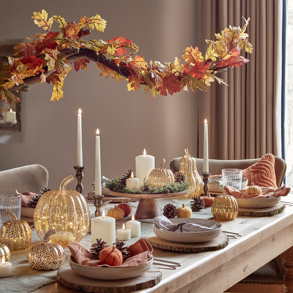 Herbstliche gedeckte Tafel mit Kerzen, Glas-Kürbissen und Wollkürbissen unter herbstlichem Blätterzweig