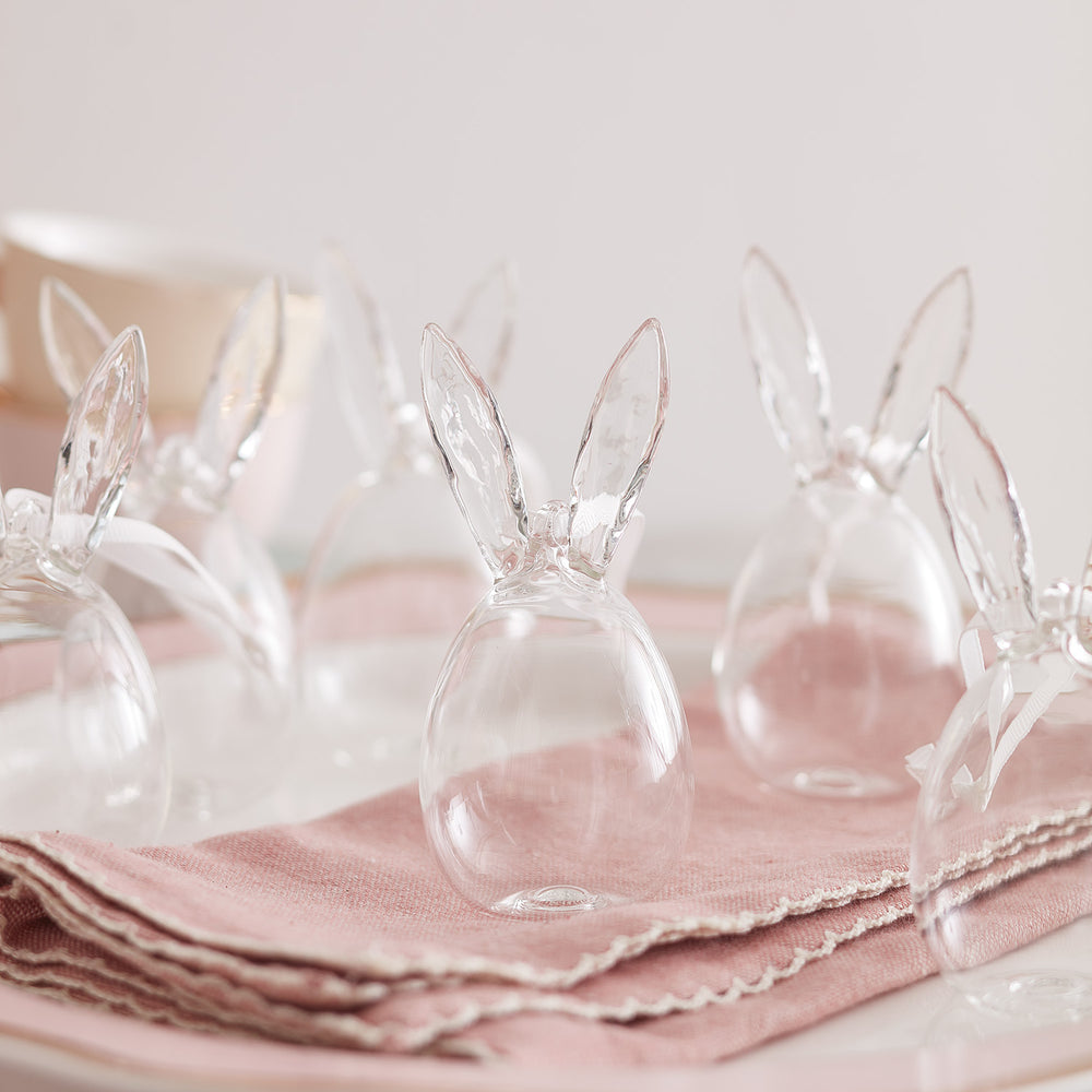 Mehrere transparente Ostereier mit Hasenohren stehen auf rosa Stoffservietten auf einem Tisch