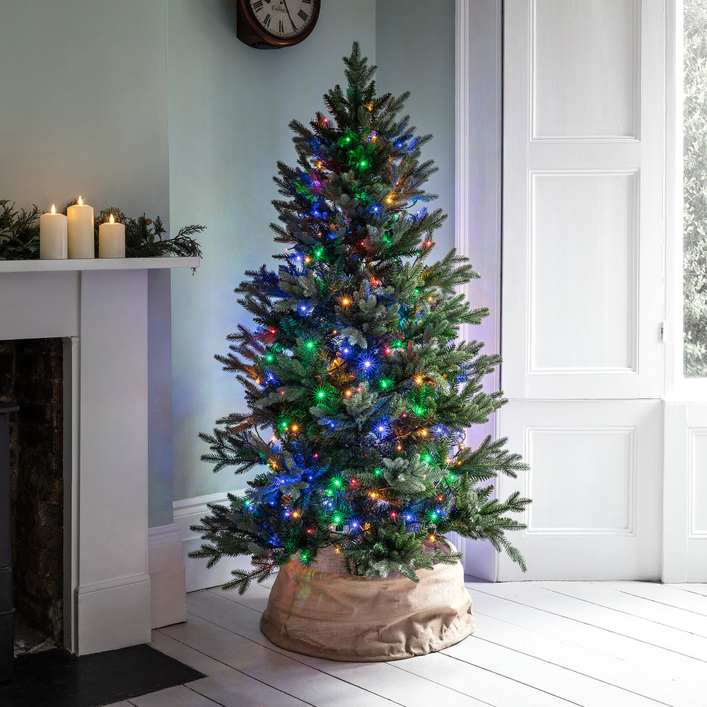 Weihnachtsbaum mit bunter LED-Lichterkette in einem hellen Raum neben Kamin und Fenster