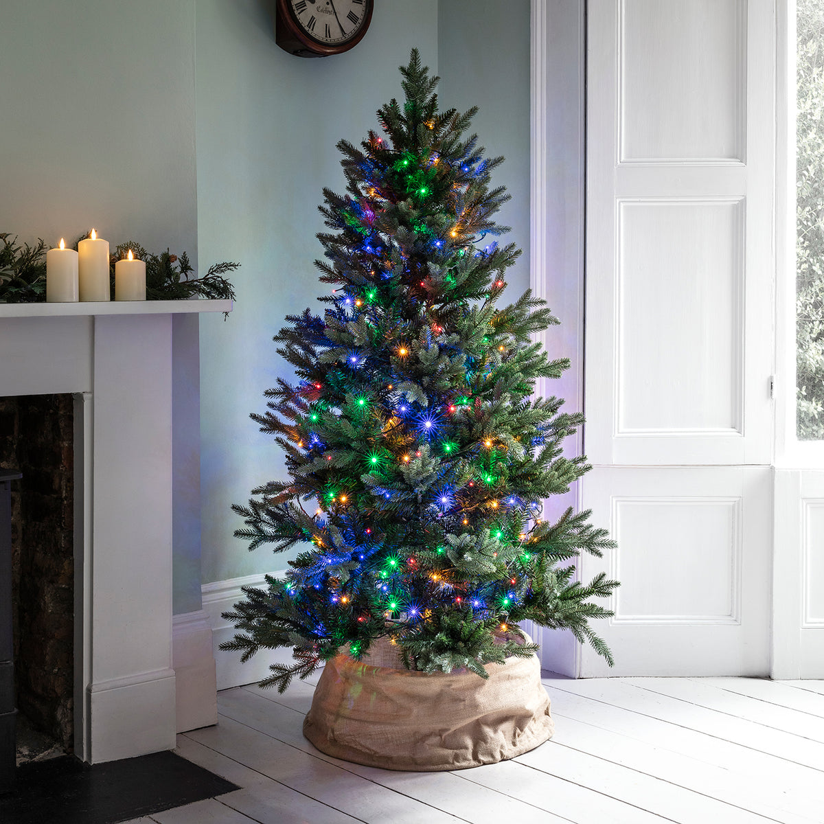 Weihnachtsbaum mit bunter LED Lichterkette in einem hellen Raum neben Kamin und Fenster