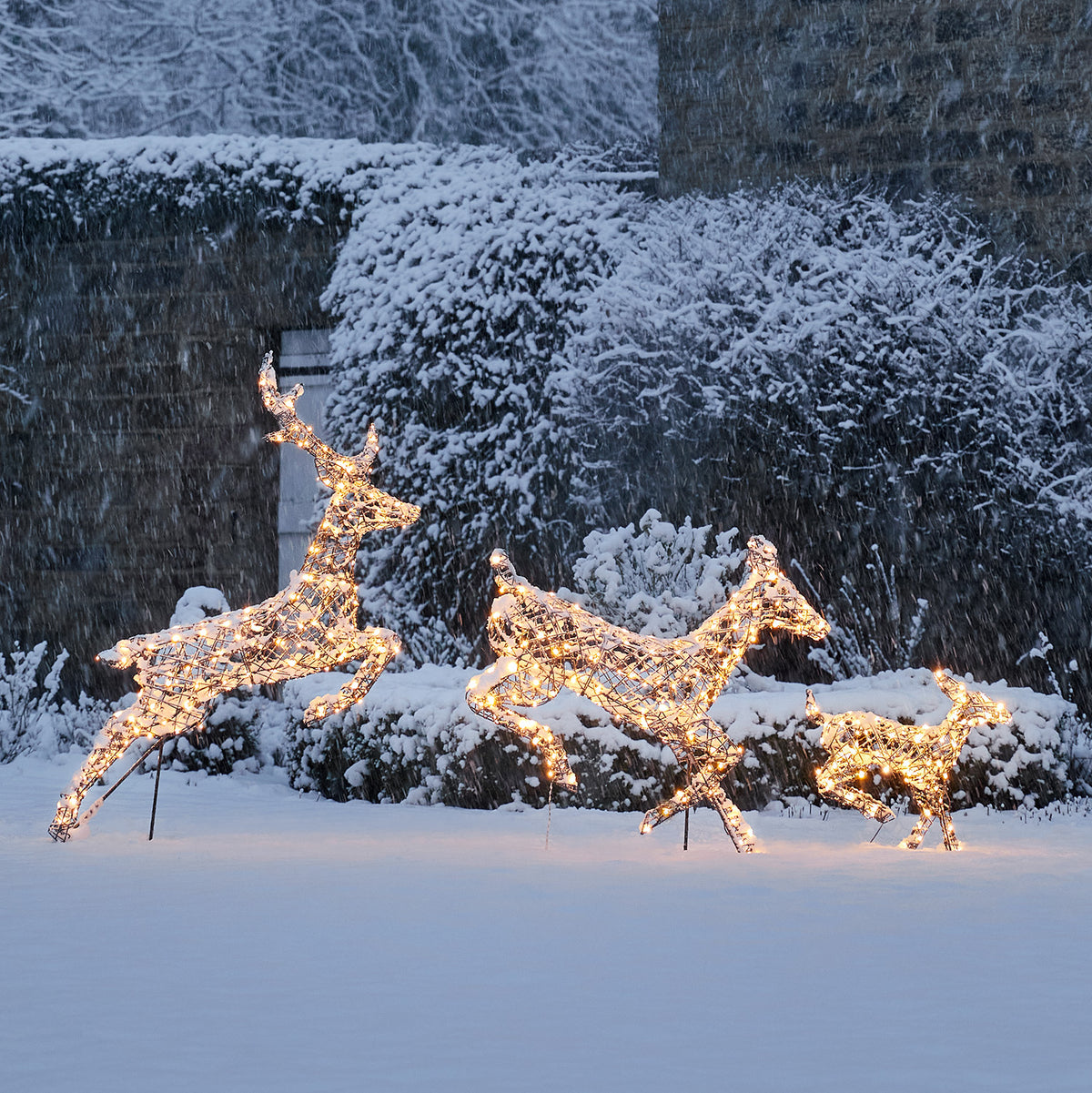Drei springende LED-Rentiere mit warmweißen Lichtern im Schnee vor Büschen