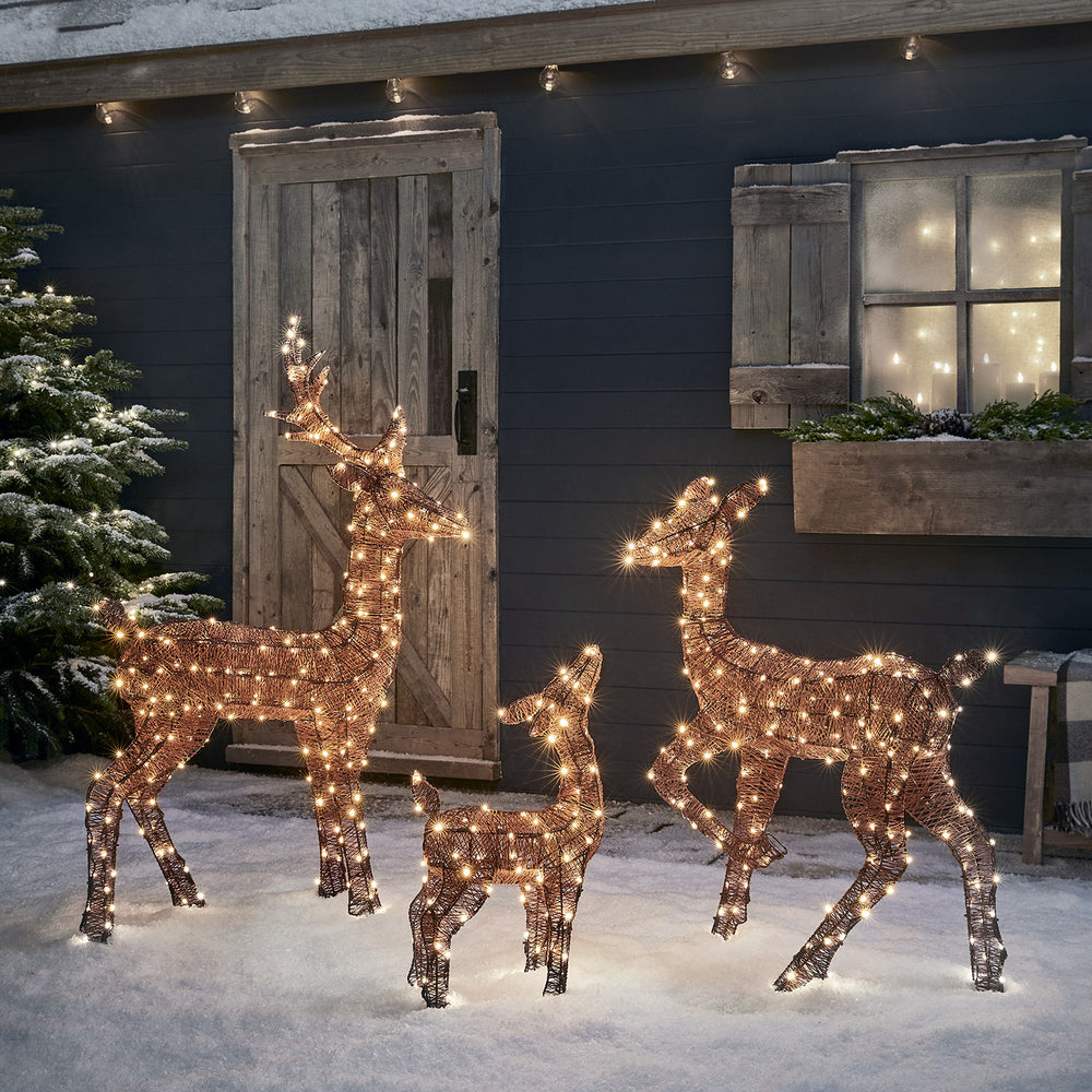 LED-Rentierfamilie mit warmweißen Lichtern vor dunkler Hauswand mit Fenster und Tür im Schnee