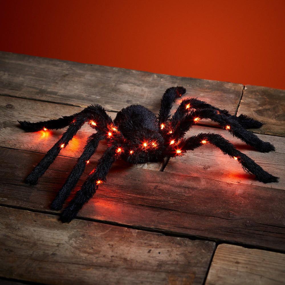 Große schwarze LED-Spinne mit 26 bernsteinfarbenen Lichtern auf Holzboden vor rotem Hintergrund