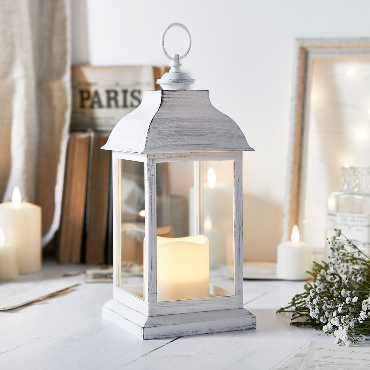 Weiße Laterne mit LED-Kerze auf weißem Holztisch, umgeben von Kerzen und Dekoration