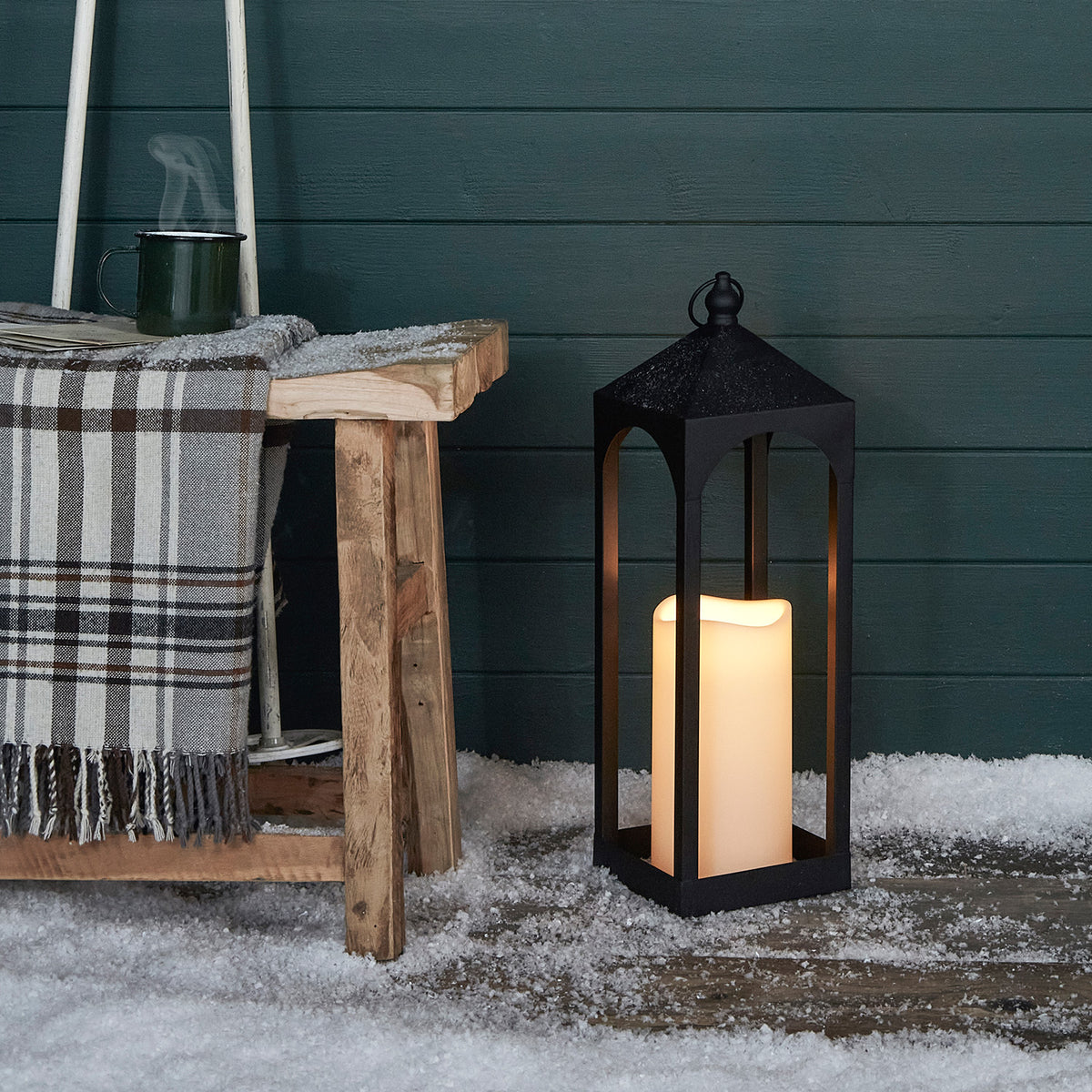 Schwarze Außenlaterne mit cremefarbener LED-Kerze steht auf schneebedecktem Boden neben Holzhocker mit kariertem Plaid