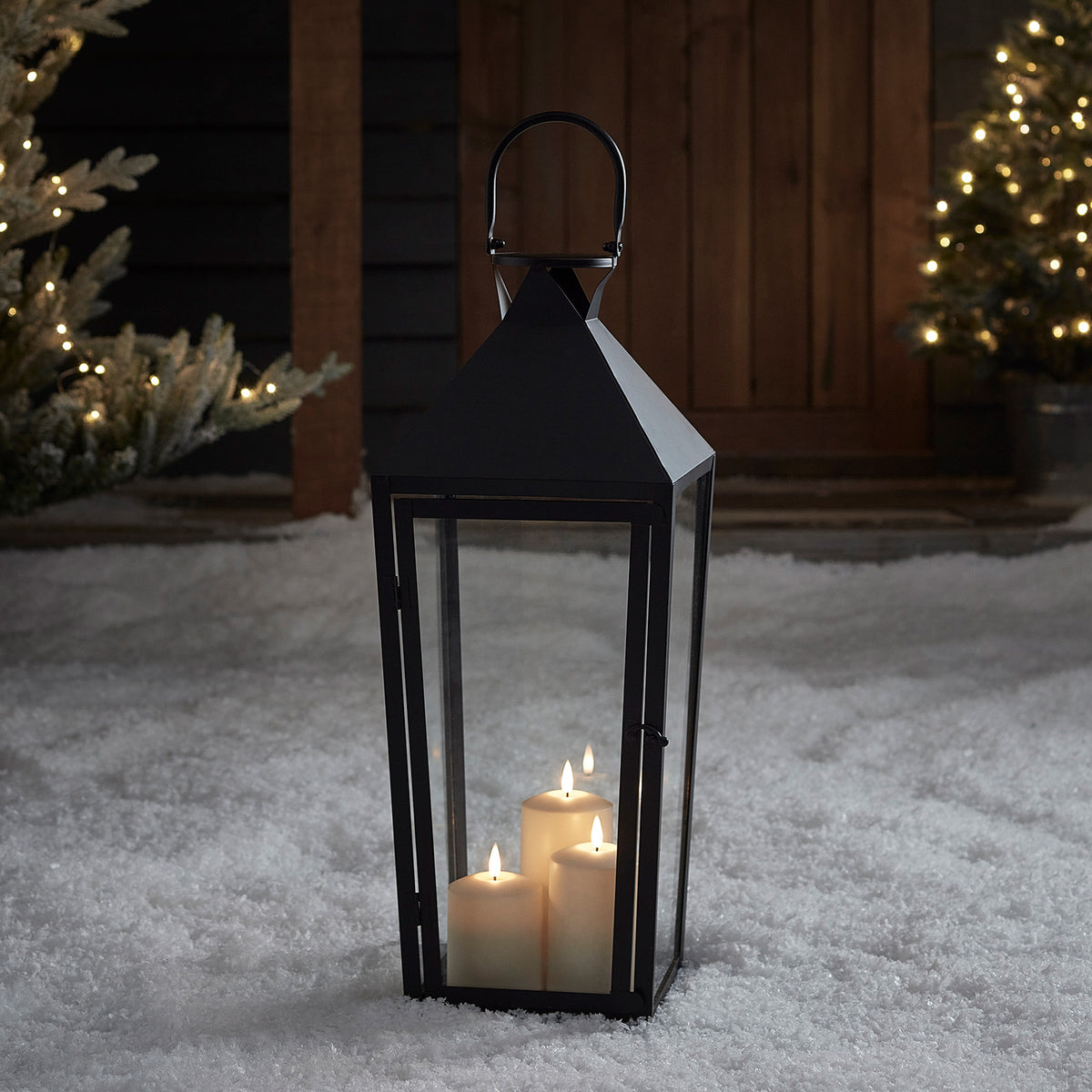 Schwarze Laterne mit drei LED-Kerzen im Schnee vor Holztür und beleuchtetem Weihnachtsbaum