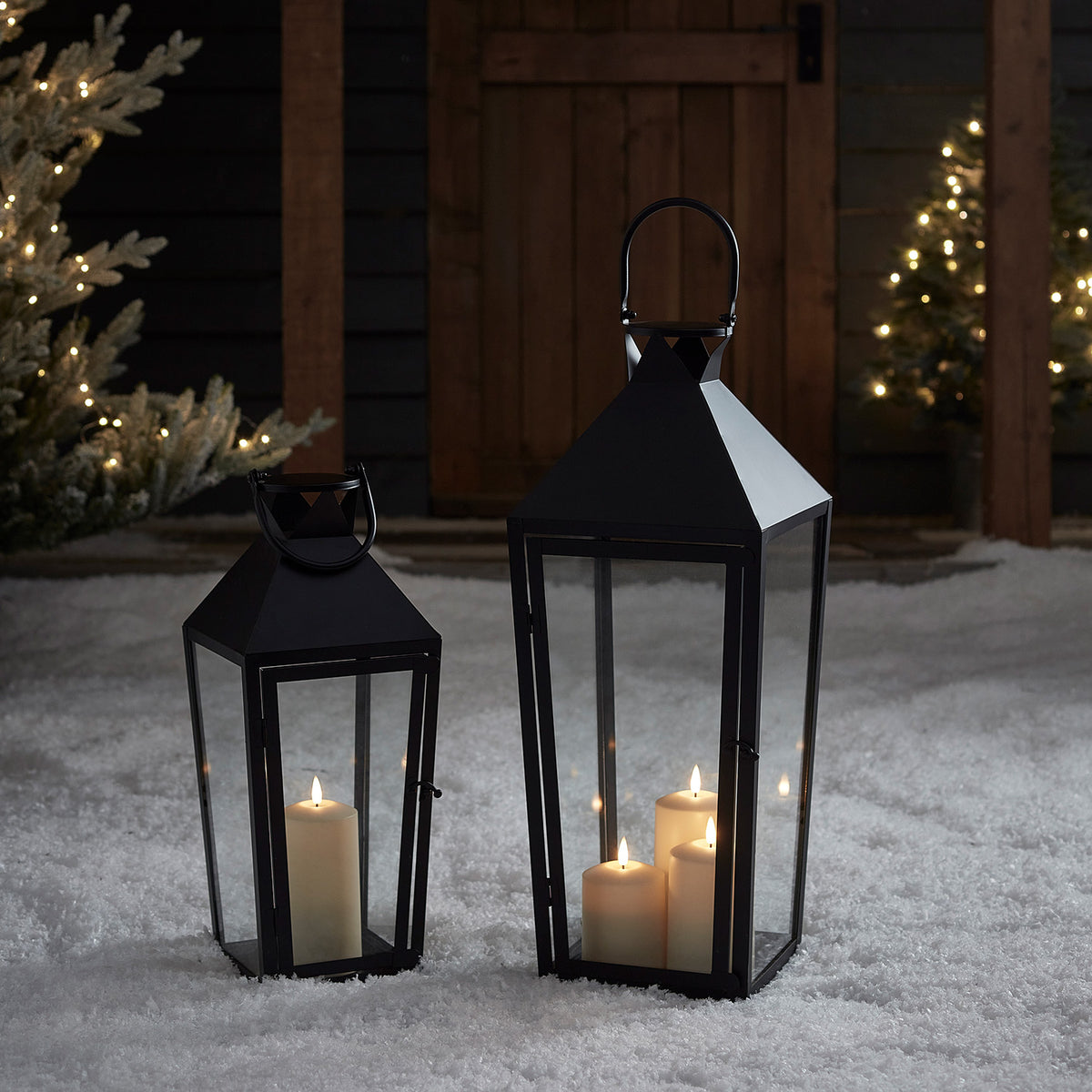 Zwei schwarze Laternen mit LED-Kerzen im Schnee vor Holztür und Weihnachtsbaum
