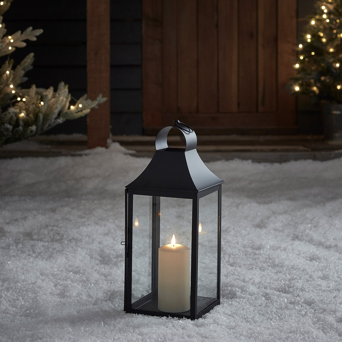 Schwarze Laterne mit LED-Kerze steht im Schnee vor dunklem Holztor und Lichterketten
