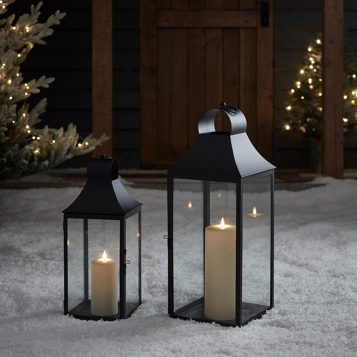 Zwei schwarze Albury Gartenlaternen mit TruGlow Kerzen im Schnee vor Holztür und Weihnachtsbeleuchtung