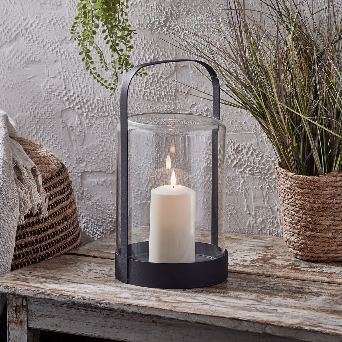 Ein schwarzes Glaswindlicht mit TruGlow® Outdoor Kerze auf einem Holztisch neben einem Korb mit Pflanzen