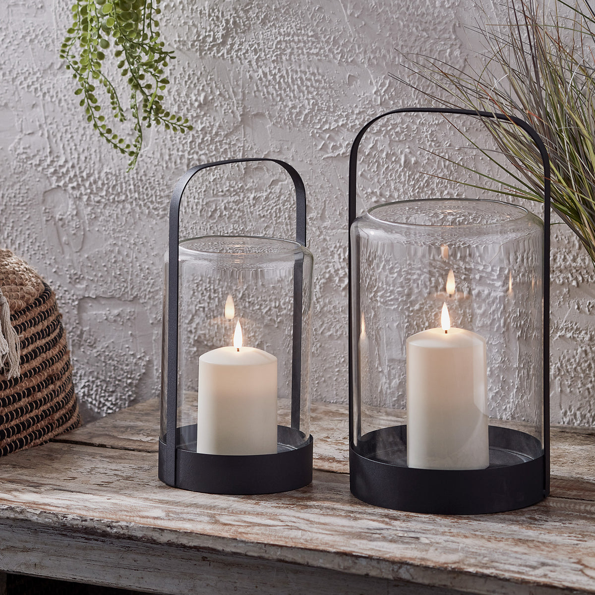 Zwei schwarze Glaswindlichter mit TruGlow® Outdoor Kerzen auf einem Holztisch vor strukturierter Wand