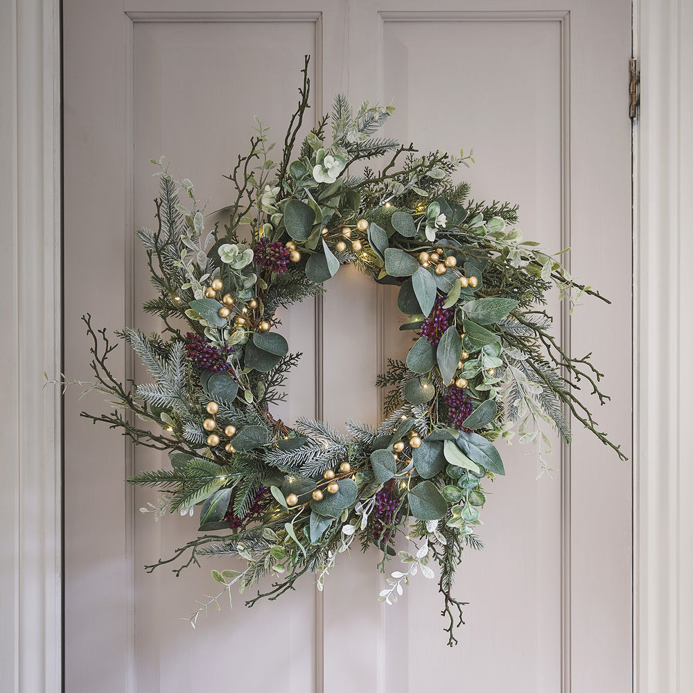 Beleuchteter Weihnachtskranz mit Eukalyptus, Kiefer, goldenen Beeren an einer hellen Tür