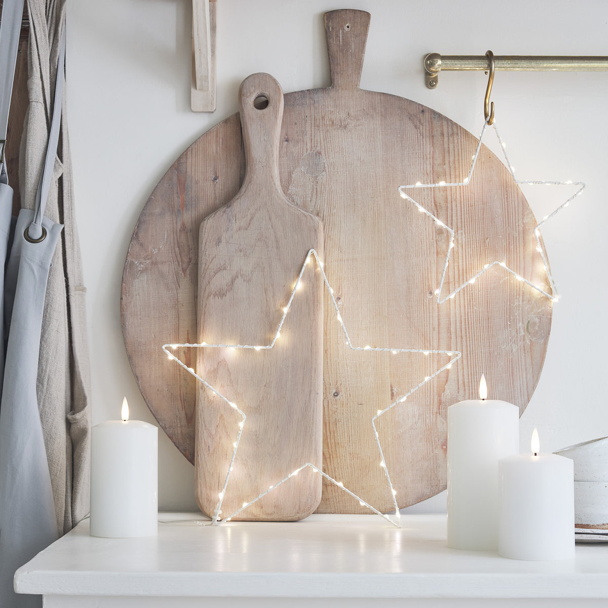 Zwei beleuchtete LED-Sterne vor Holzbrett und drei weiße LED-Kerzen auf weißem Tisch