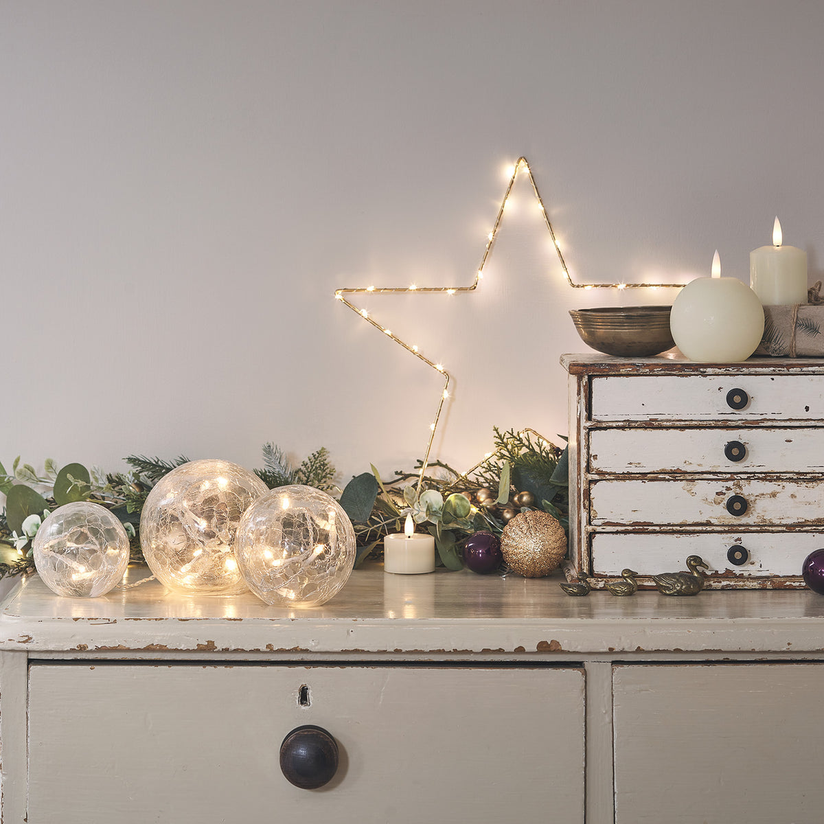 LED-Glaskugeln auf einem weißen Sideboard mit Stern aus Lichterkette an der Wand
