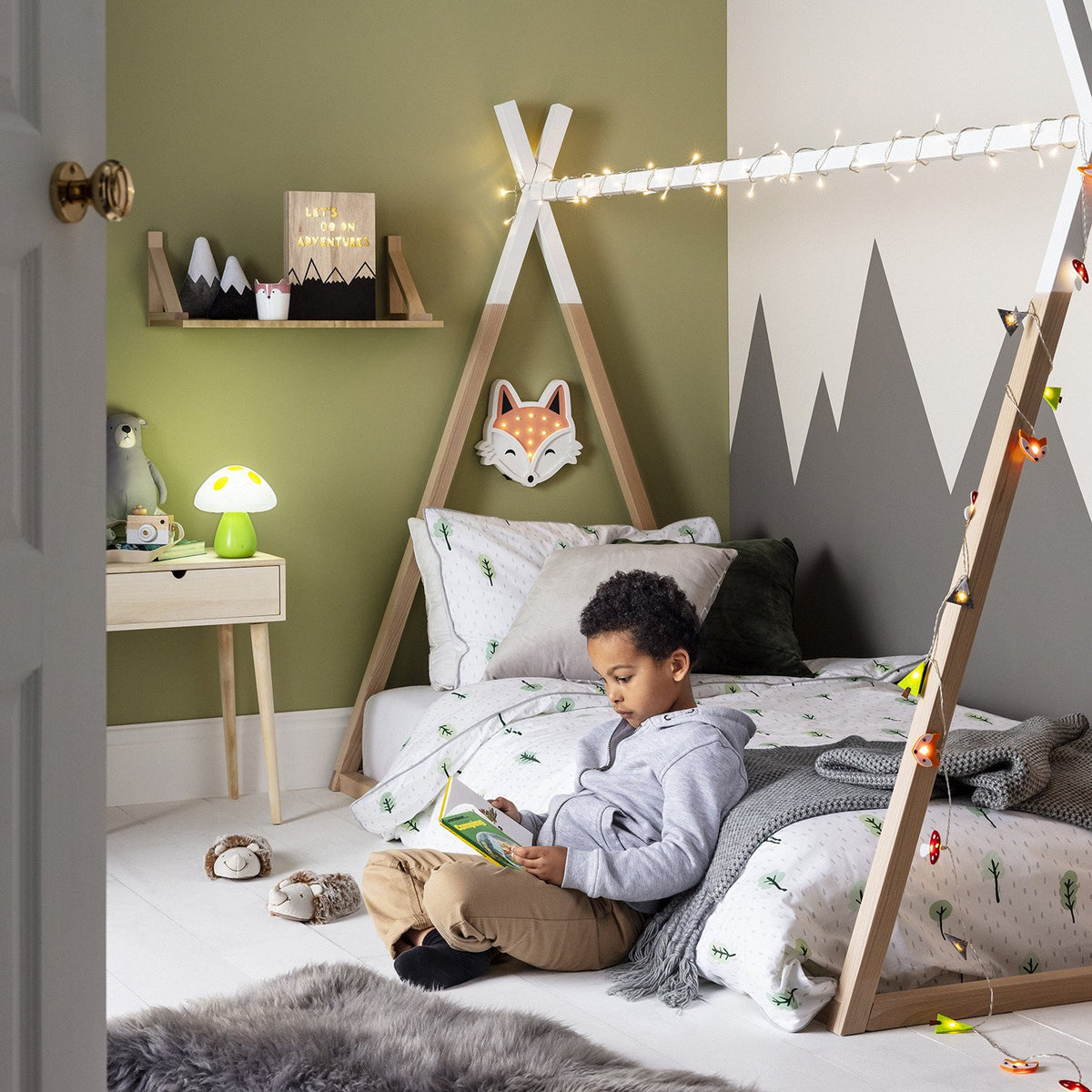 Kinderzimmer mit Tipi-Bett, Lichterkette mit Waldmotiven und Kind liest Buch auf dem Bett