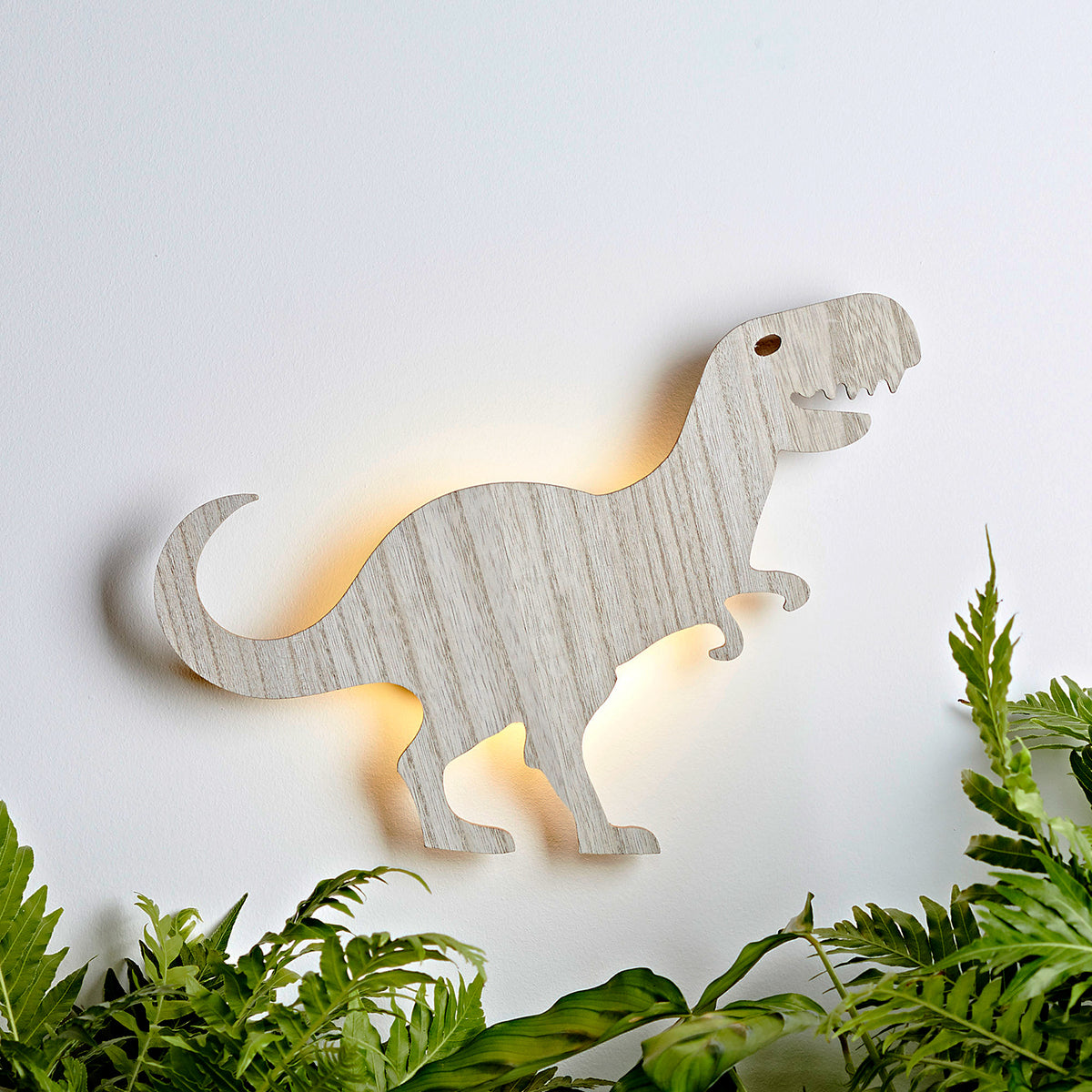 Holz-Wandlampe in Form eines T-Rex mit warmem Licht an weißer Wand, umgeben von grünen Pflanzen