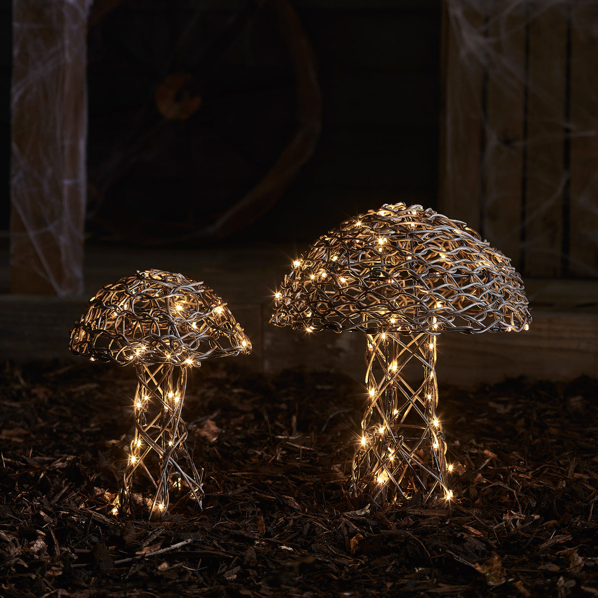 Zwei leuchtende Pilzfiguren aus Rattan mit warmweißen LEDs im dunklen Garten auf Mulchboden