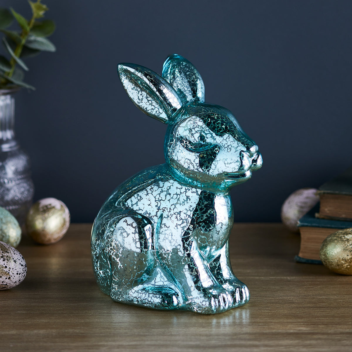 Blauer Glas-Hase mit Melierung und LEDs auf Holztisch vor dunklem Hintergrund mit Osterdekoration
