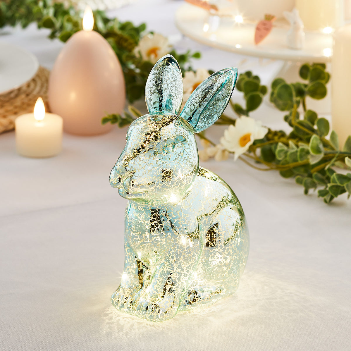 Blauer Glas-Hase mit warmweißen LEDs auf hellem Tisch mit Kerzen und Deko im Hintergrund