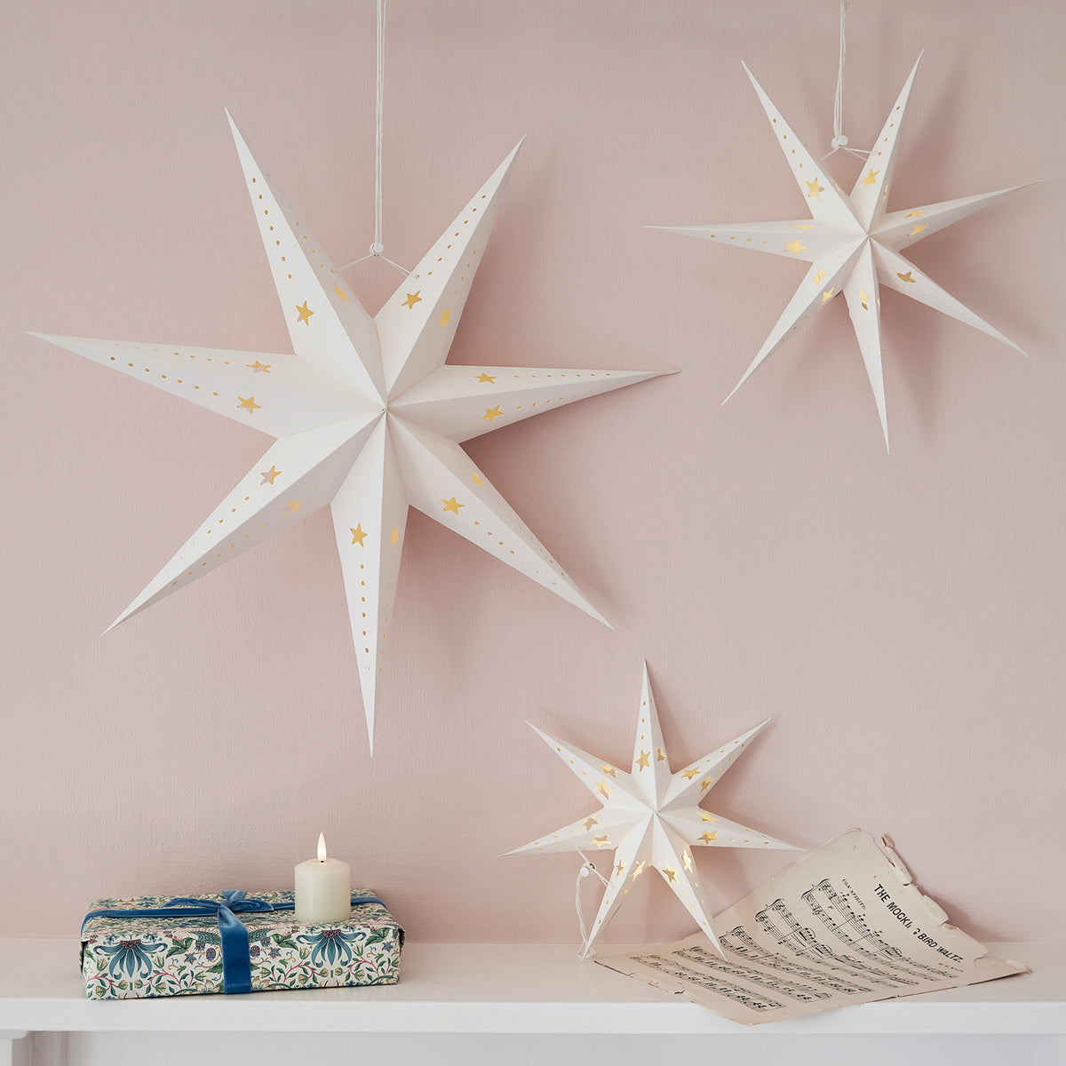 Drei weiße Papiersterne mit LED-Licht hängen vor einer rosa Wand über einem Tisch mit Geschenk und Notenblatt