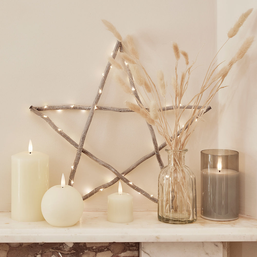 Beleuchteter Holzstern mit warmweißen LEDs auf Kaminsims mit Kerzen und Vase mit Trockenblumen