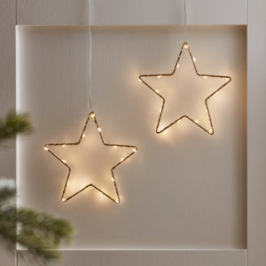 Zwei goldene LED-Sterne hängen an dünnen Kabeln vor einer hellen Wand