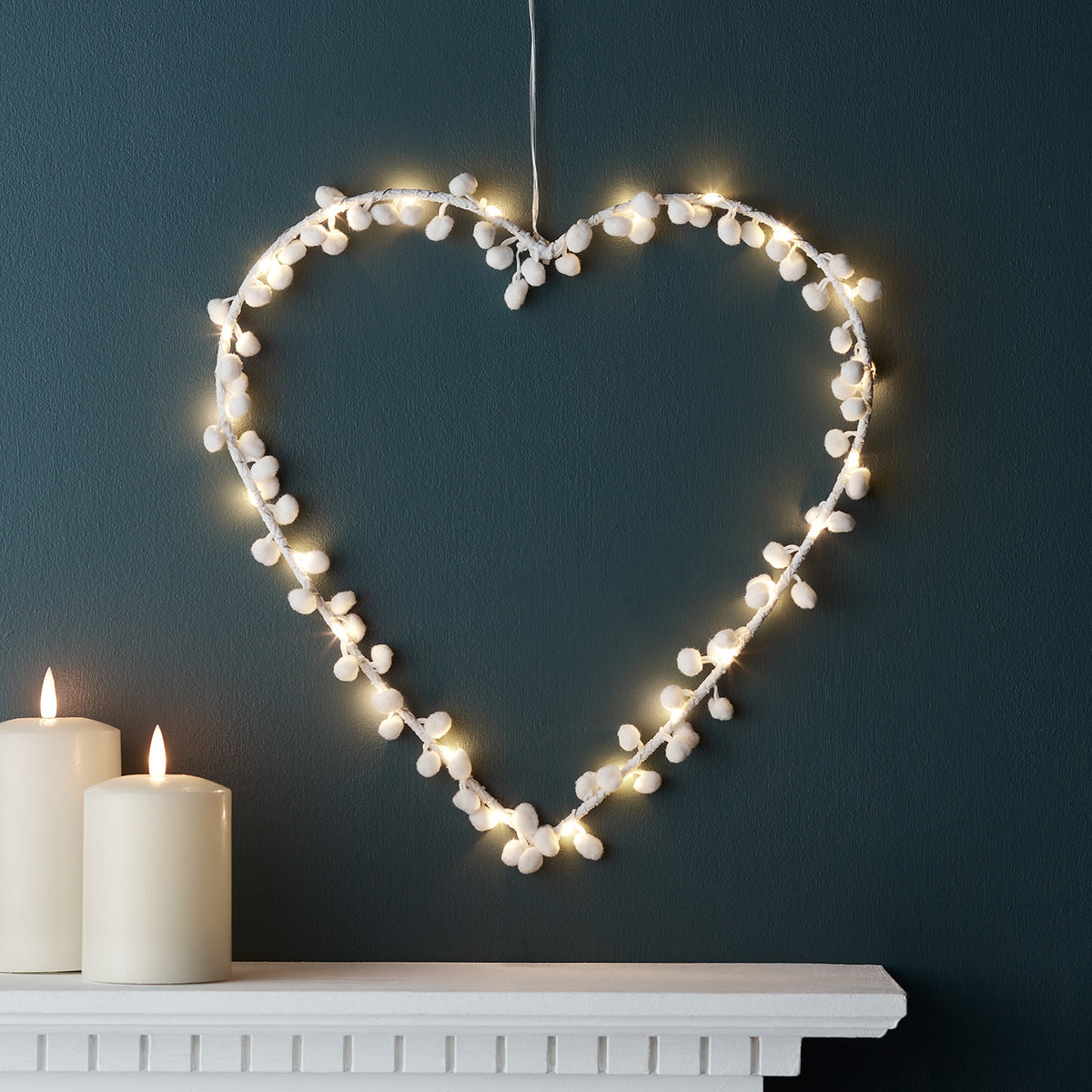 Beleuchtetes Micro LED Herz mit weißen Pompons an dunkler Wand über Kaminsims mit Kerzen