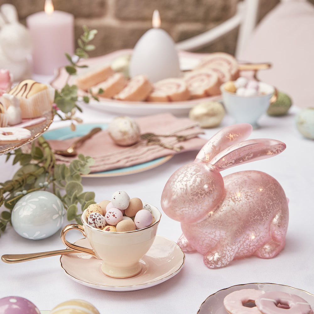 Pastellrosa Glashase mit LED neben Tasse mit bunten Ostereiern und Ostergebäck