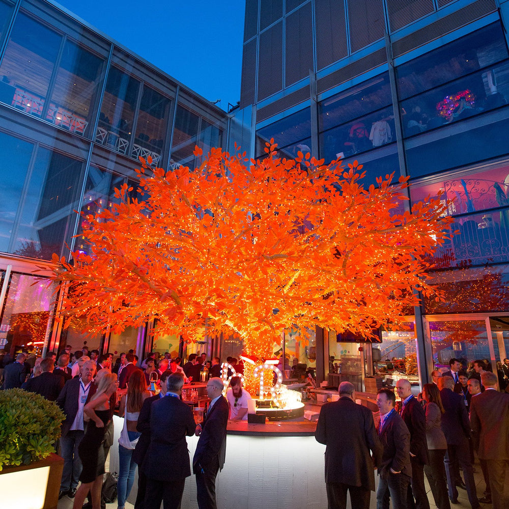Orangene LED-Lichterkette um Baumstamm bei Abendveranstaltung mit Gästen im Hintergrund
