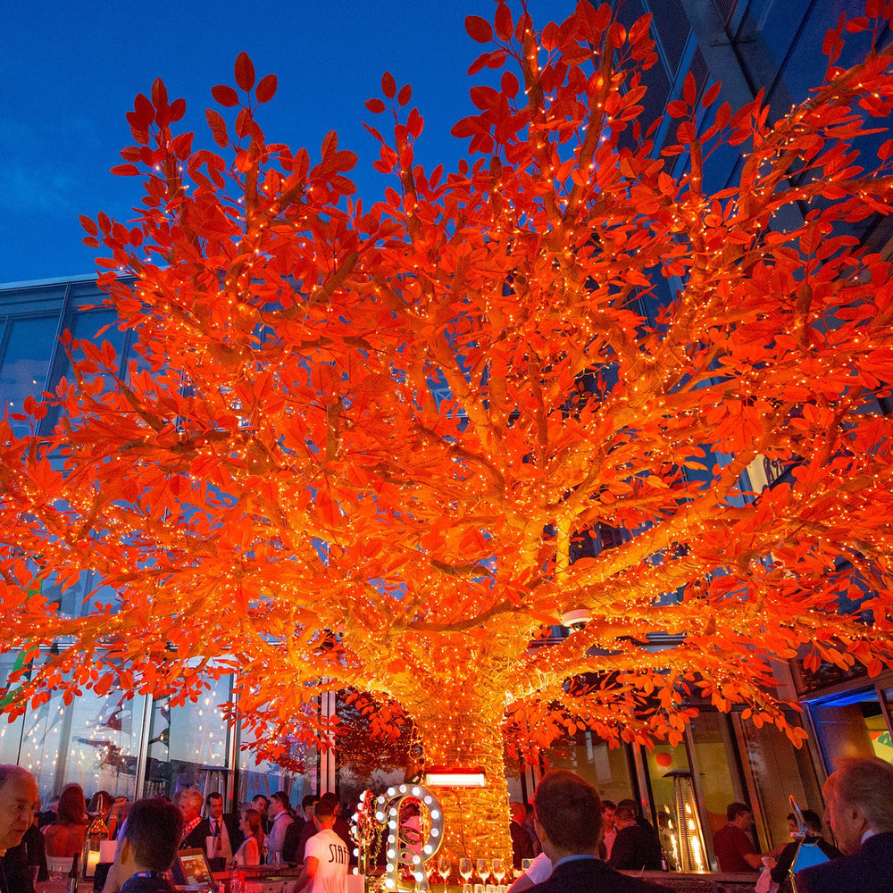 Orangener Baum mit leuchtenden LED-Lichterketten vor blauem Abendhimmel und Menschen im Hintergrund