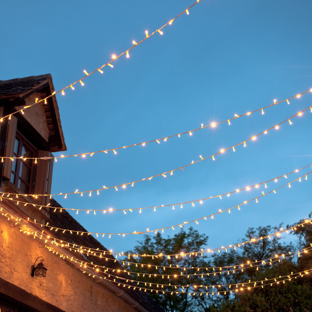 Mehrere Reihen warmweißer LED-Lichterketten mit transparentem Kabel über einem Außenbereich bei Abenddämmerung