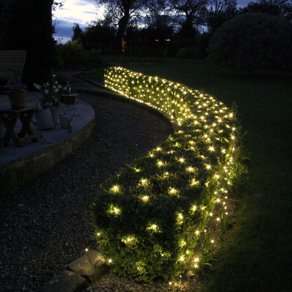 Warmweiße LED-Lichterkette dekoriert Hecke im Garten bei Dämmerung