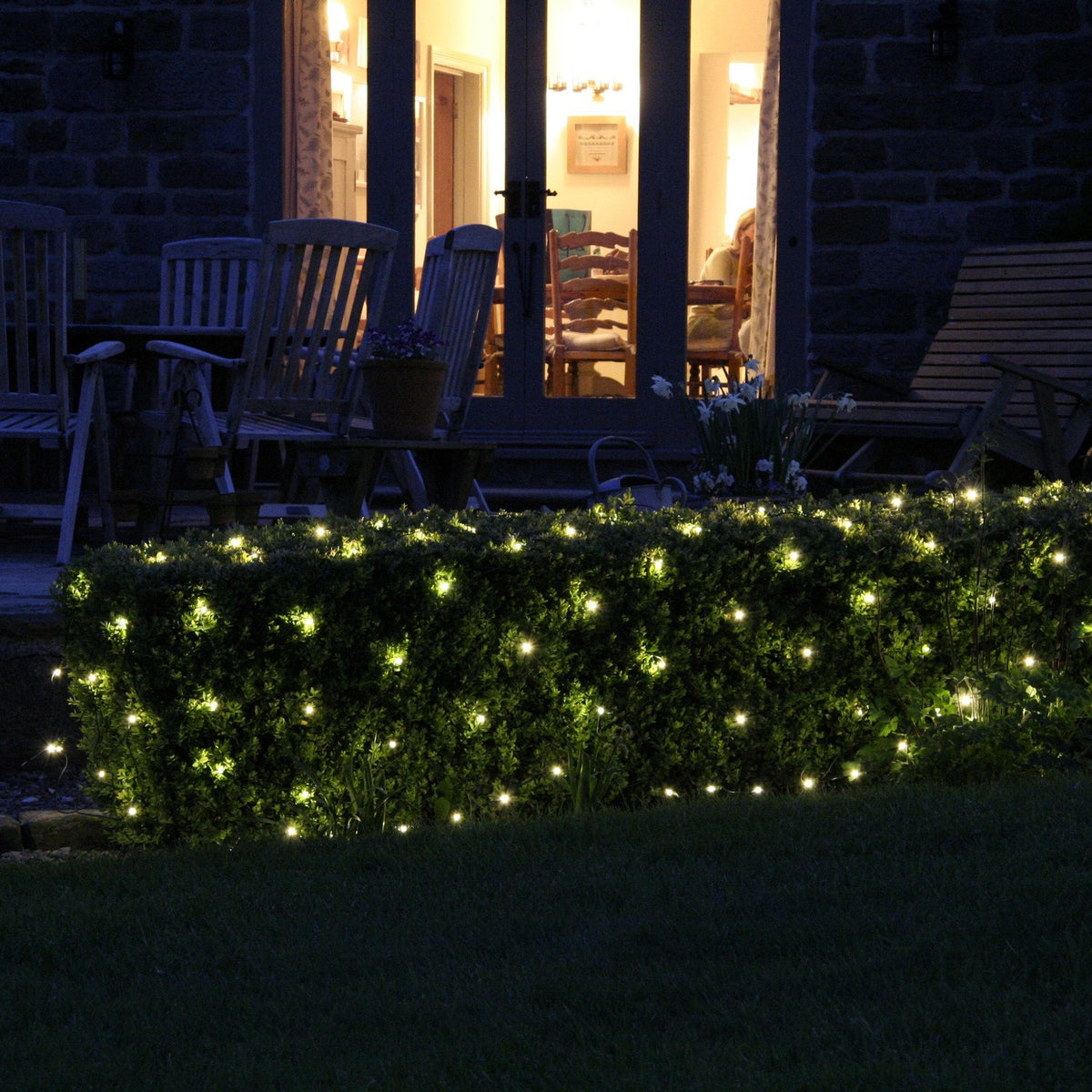 Warmweiße LED-Lichterkette beleuchtet Hecke vor Terrasse mit Gartenmöbeln