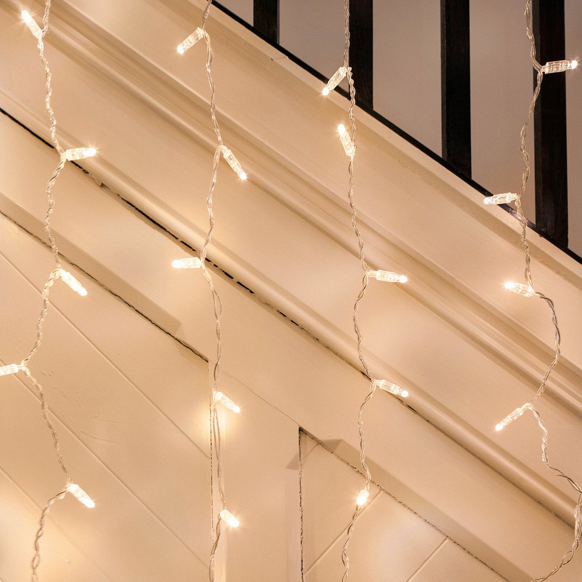 Warmweiße LED-Lichtervorhänge mit transparentem Kabel hängen an einer weißen Wand und Treppe