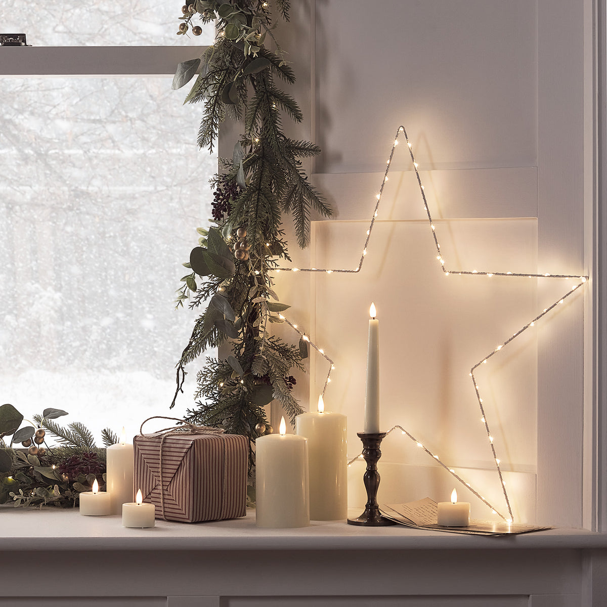 Weißer LED-Stern mit warmweißen Lichtern an Wand neben Kerzen, Geschenk und Tannenzweigen