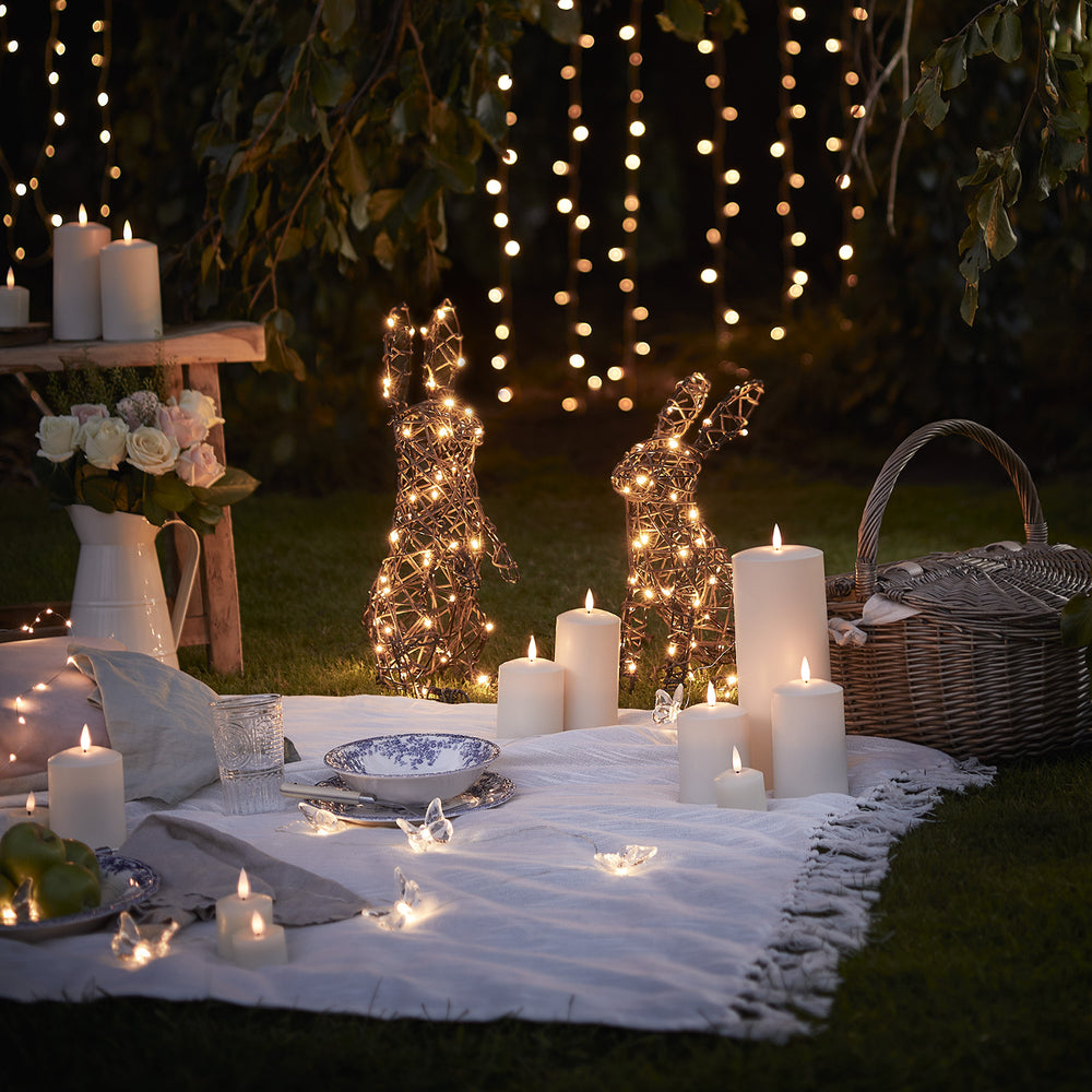 Beleuchtete LED Hasen aus Rattan neben Kerzen und Picknickdecke im Garten bei Abendlicht