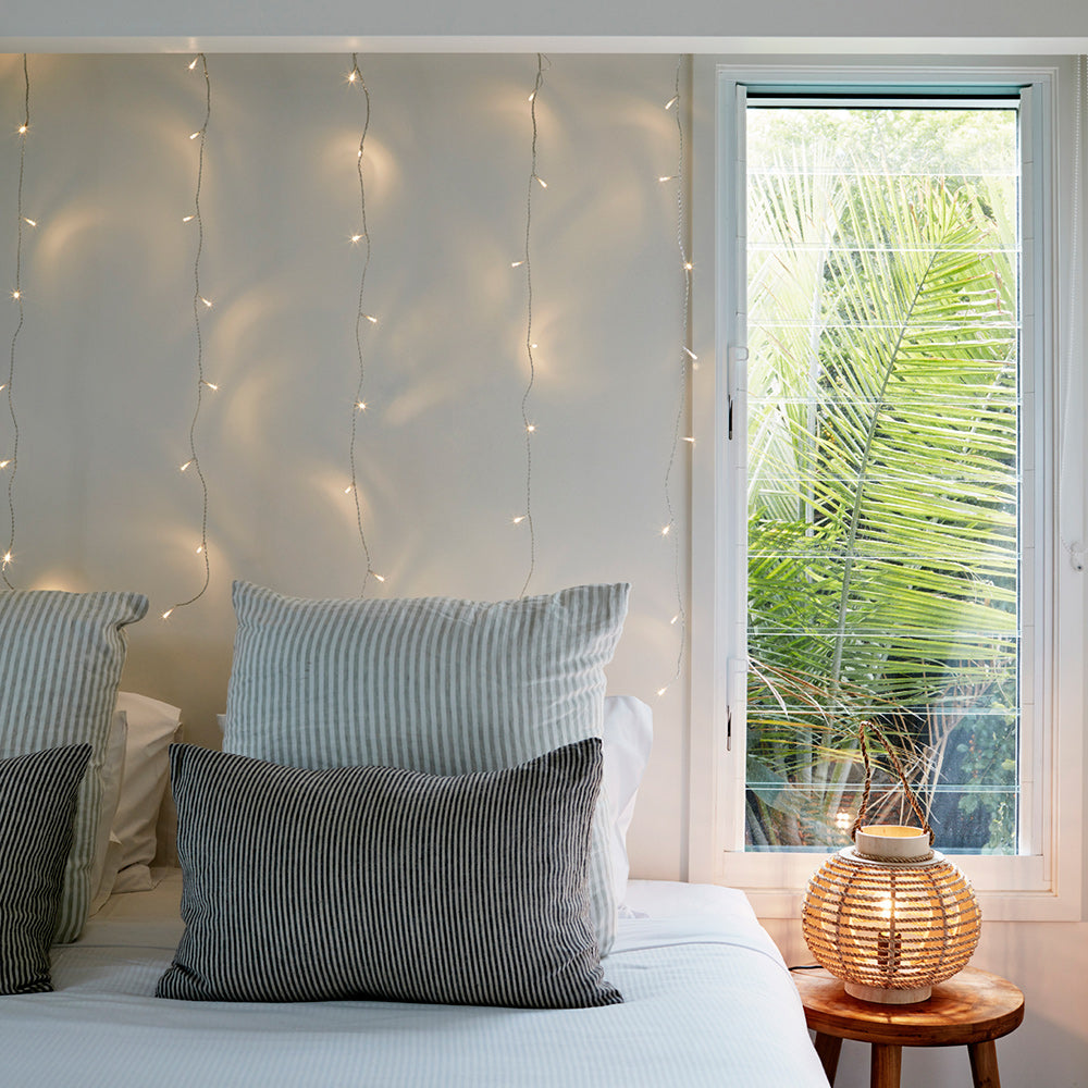 Schlafzimmer mit warmweißen LED-Lichterketten an der Wand, Bett mit Kissen und Fenster mit Blick auf grüne Pflanzen