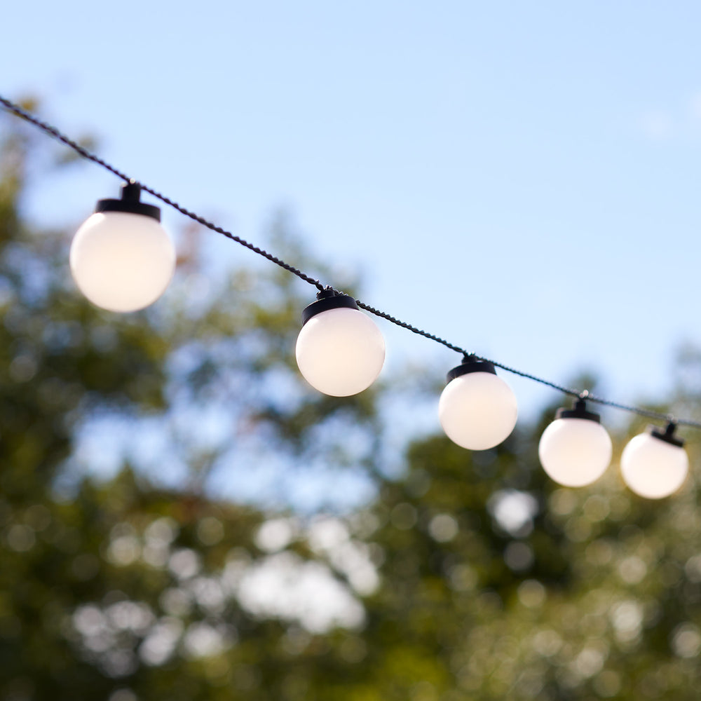 Lichterkette mit weißen Kugellampen vor blauem Himmel und unscharfen Bäumen im Hintergrund