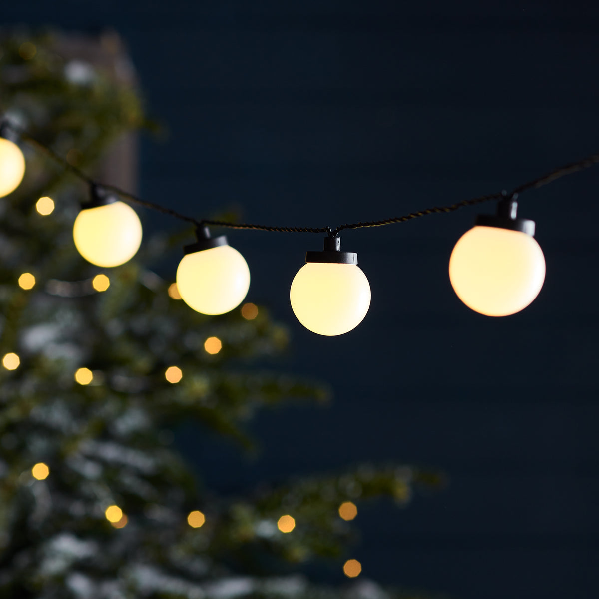 Beleuchtete Lichterkette mit weißen Kugellampen vor dunklem Nachthimmel und unscharfen Lichtern