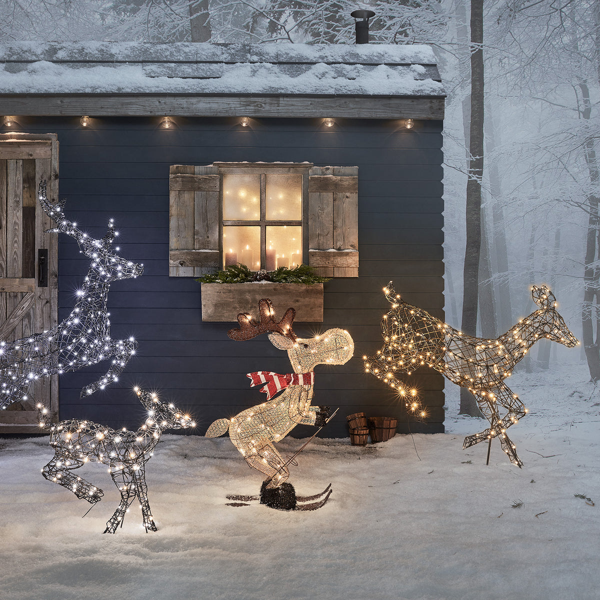 Drei beleuchtete Weihnachtsfiguren, darunter Elch auf Skiern vor dunklem Haus mit Schnee