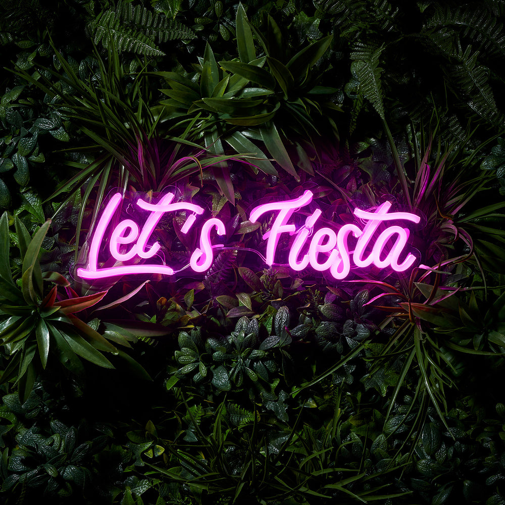 Leuchtender pinkfarbener LED-Neonschriftzug Let's Fiesta vor dunklem Pflanzenhintergrund