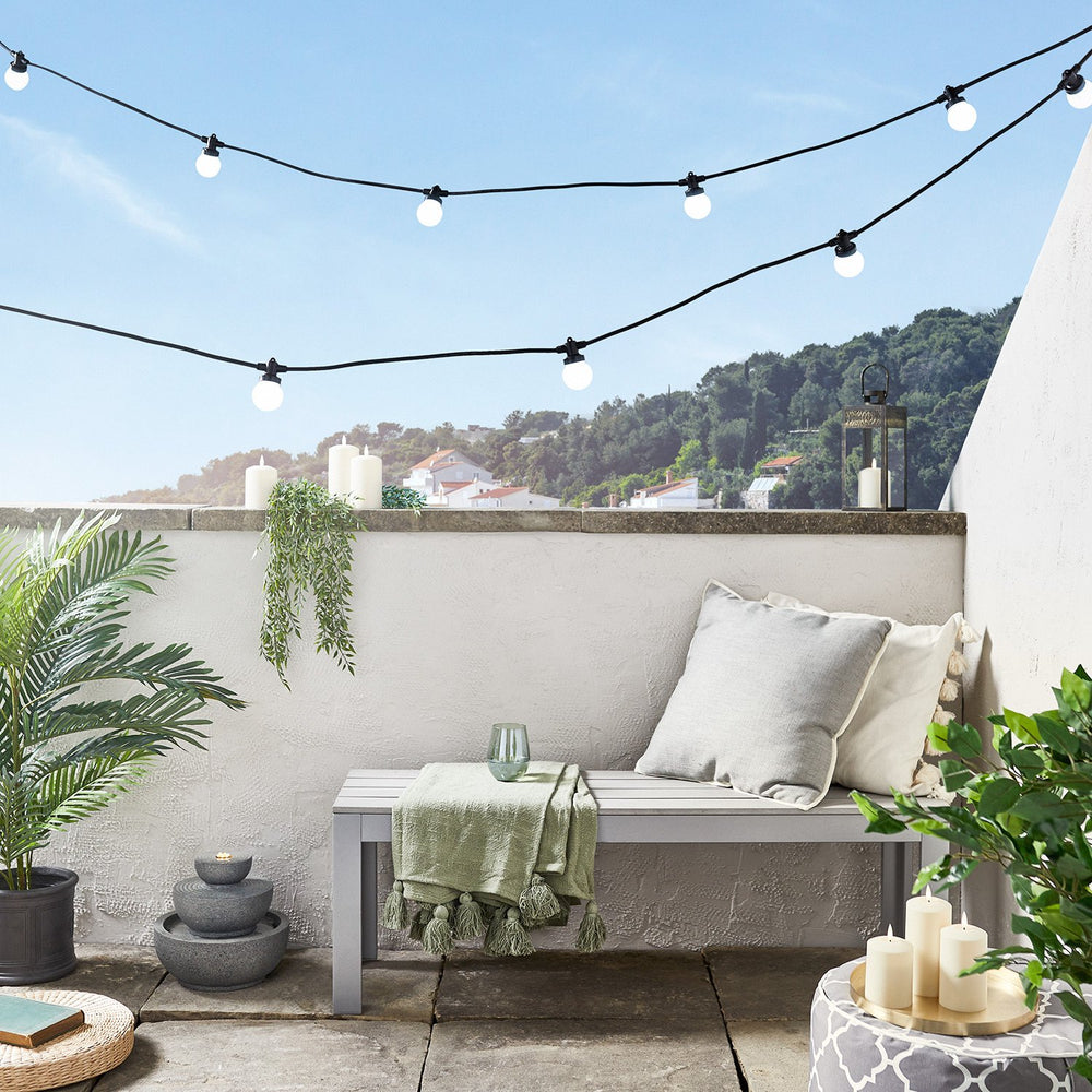 Zwei Reihen weiße LED-Glühbirnen an schwarzem Kabel über Terrasse mit Bank und Pflanzen