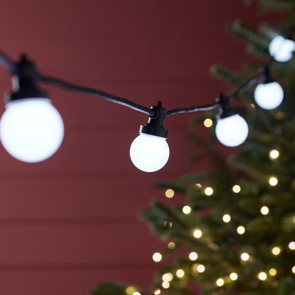 Weiße LED-Kugellichter an schwarzem Kabel vor roter Wand und Weihnachtsbaum