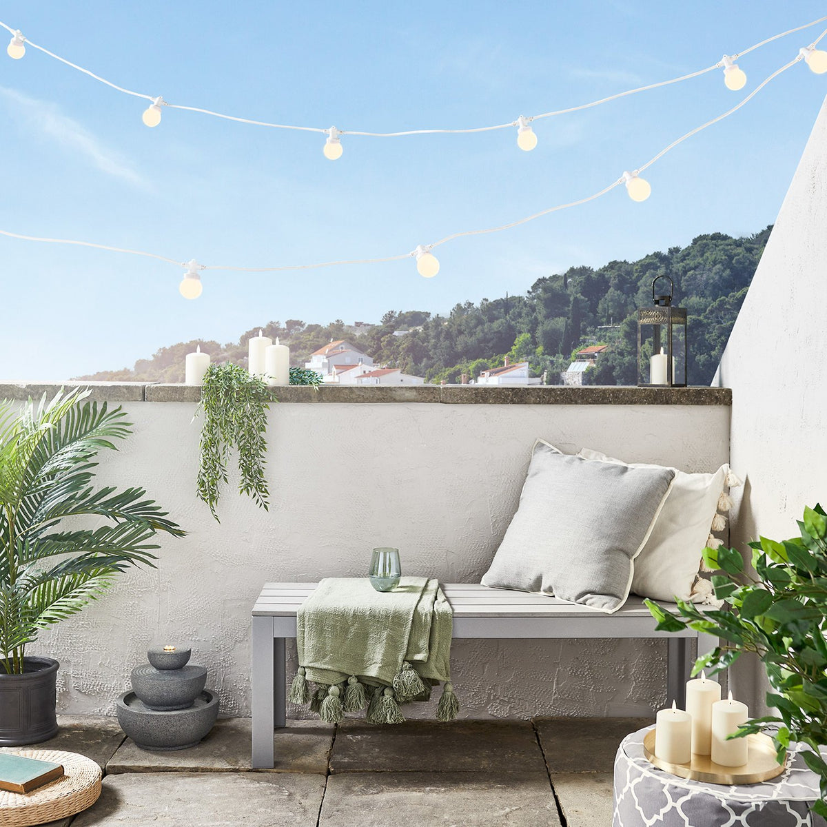 Weiße Lichterkette mit warmweißen Glühbirnen über Sitzbank auf Balkon mit Pflanzen und Kerzen